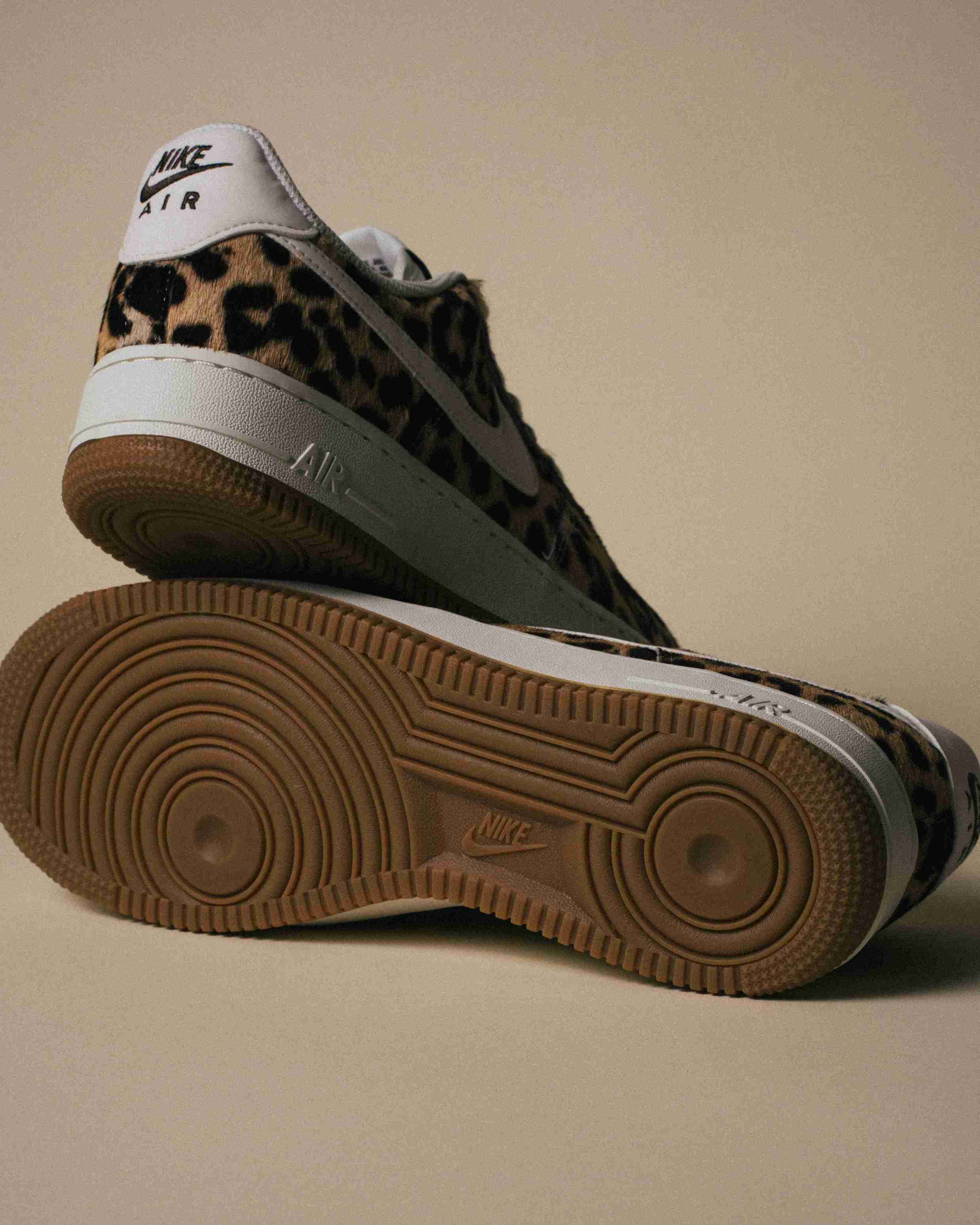 Paire de sneakers Nike Air Force 1 à motif léopard, placée à côté d’une boîte Nike grise sur un fond beige, vue artistique avec lumière douce.